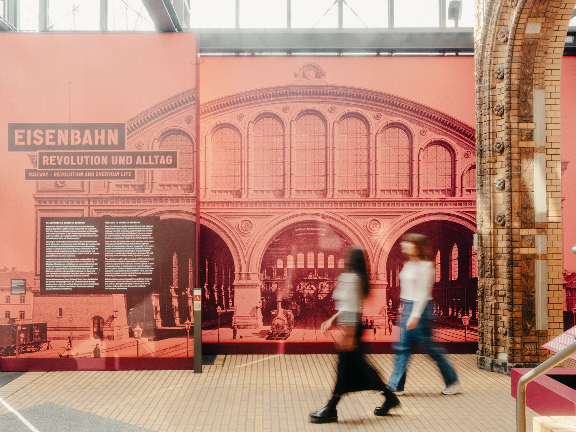 Ausstellung „Eisenbahn – Revolution und Alltag“ im Deutschen Technikmuseum. Eine großformatige historische Bahnhofsfassade in Rot-Tönen, davor gehen zwei Besucherinnen vorbei.