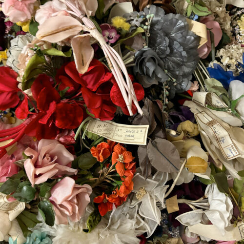 Ein Haufen künstlicher Blumen in verschiedenen Farben und Formen, teils mit Etiketten versehen, die Beschriftungen wie „R.E. Flowers“ tragen. Die bunte Sammlung umfasst rote, rosa, graue und weiße Blüten.