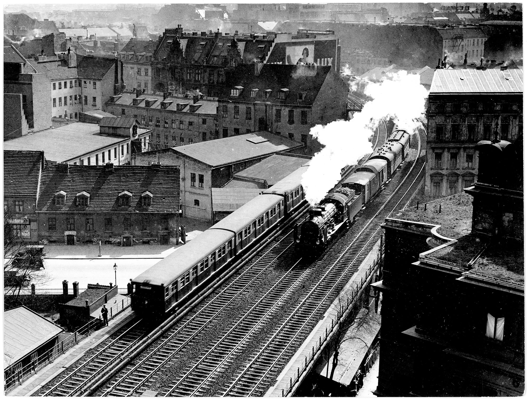 Historische Stadtansicht mit einer dampfenden Dampflokomotive auf einer erhöhten Eisenbahnstrecke. Im Vordergrund Wohnhäuser und Gebäude, im Hintergrund eine Stadtlandschaft mit dicht stehenden Häusern.