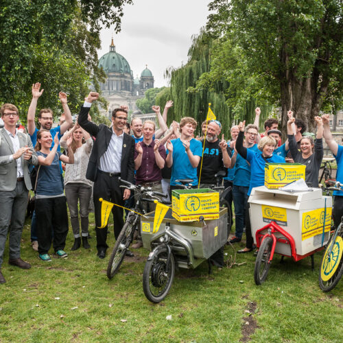 Gruppe von Menschen jubelt im Freien neben Fahrrädern und Lastenrädern mit gelben Boxen, die mit „Volksentscheid Fahrrad“ beschriftet sind, vor einer grünen Kulisse und der Kuppel des Berliner Doms im Hintergrund.