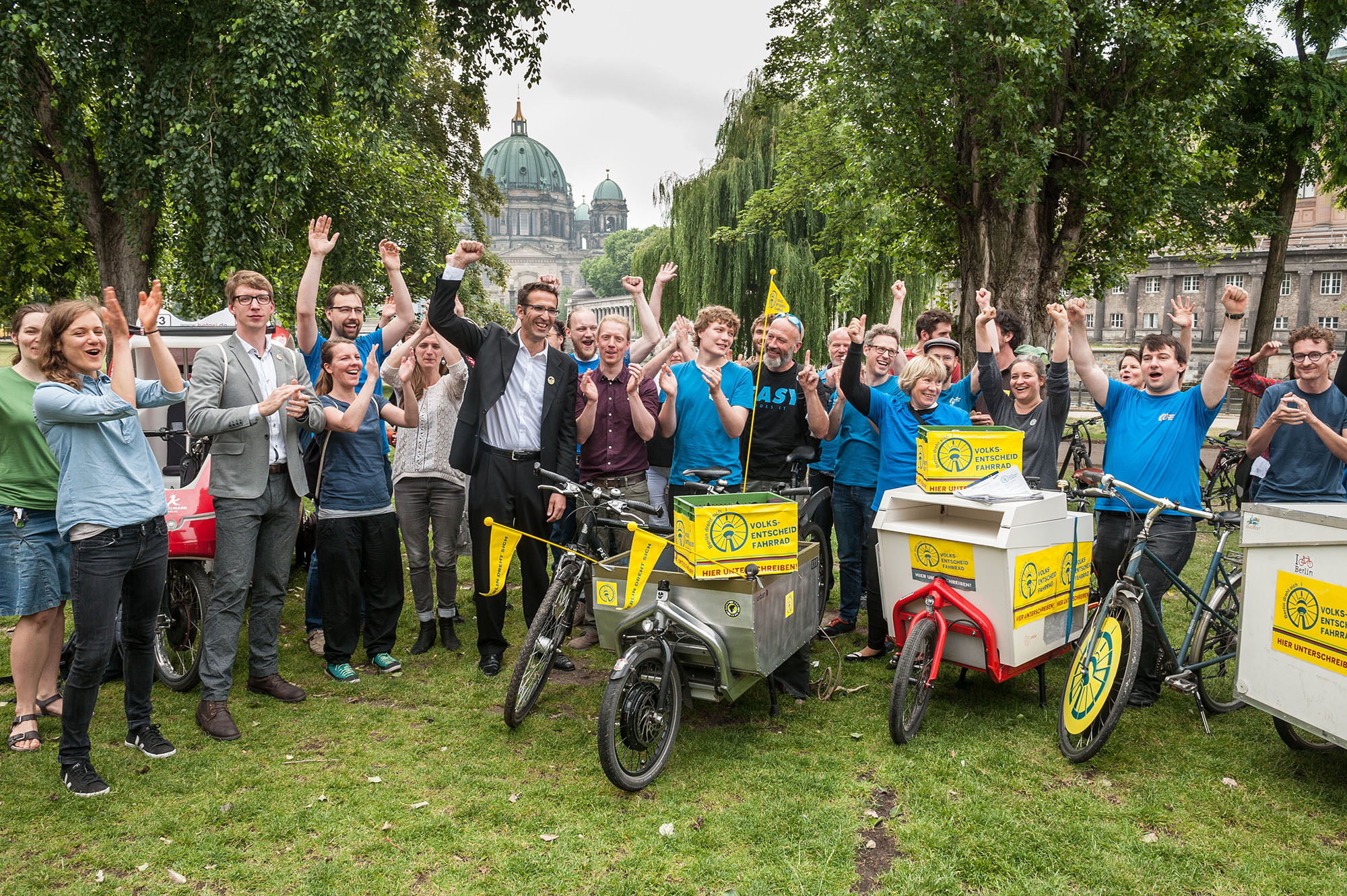 Gruppe von Menschen jubelt im Freien neben Fahrrädern und Lastenrädern mit gelben Boxen, die mit „Volksentscheid Fahrrad“ beschriftet sind, vor einer grünen Kulisse und der Kuppel des Berliner Doms im Hintergrund.