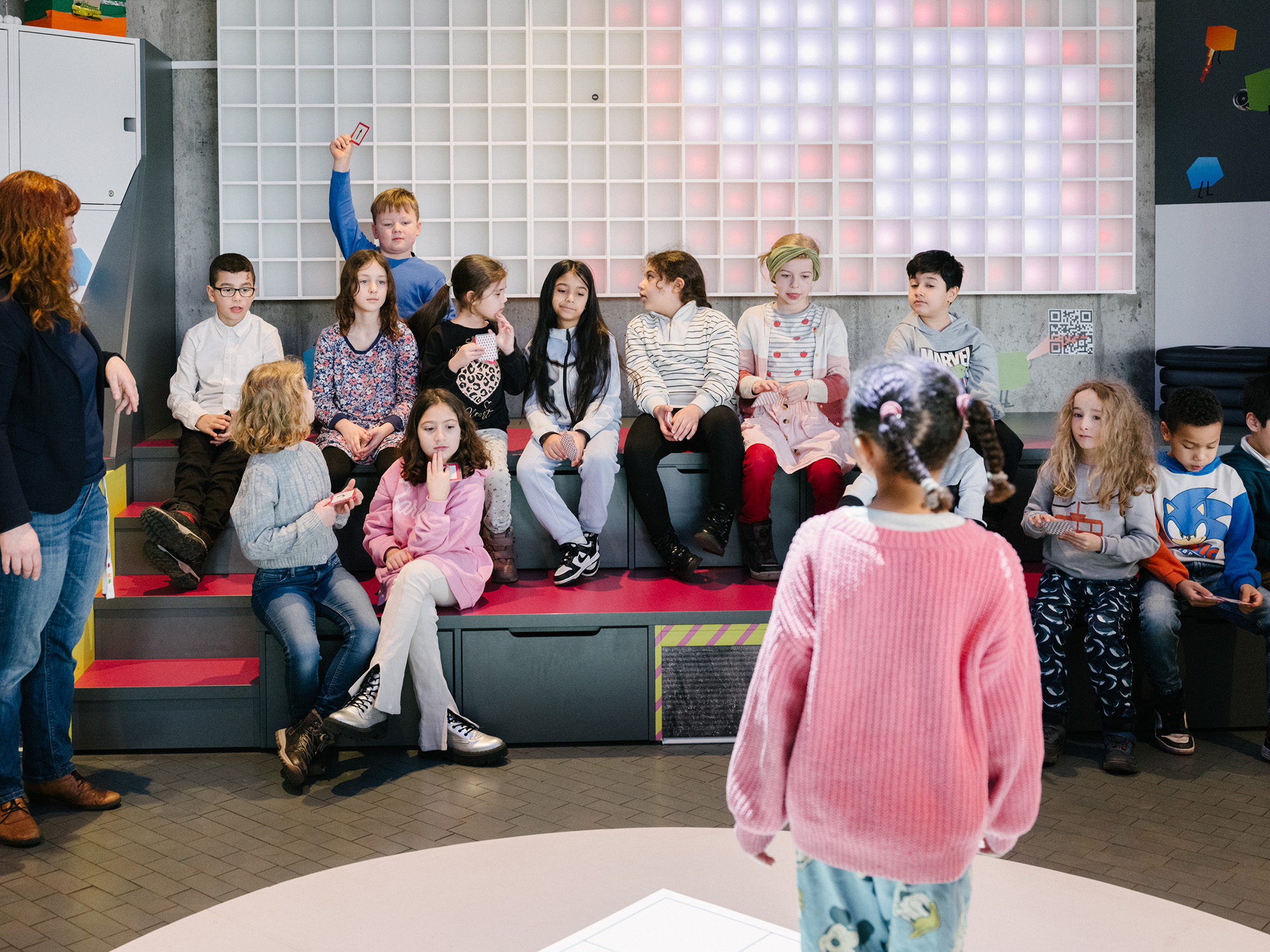 Eine Gruppe von Kindern sitzt auf bunten Sitzstufen in einem modernen Raum mit einer beleuchteten Pixelwand. Ein Mädchen im rosa Pullover steht vorne, während eine Frau links die Kinder betreut.