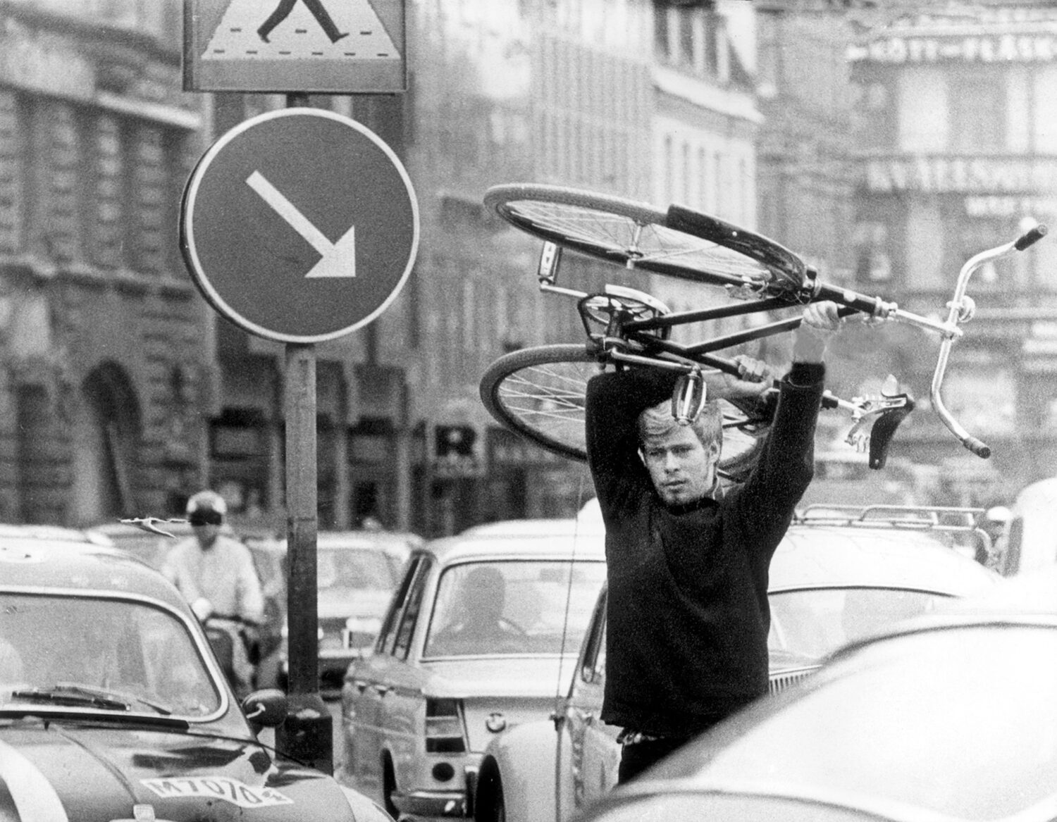 Ein Mann trägt ein Fahrrad über seinem Kopf, während er sich durch dichten Verkehr bewegt. Im Hintergrund sind Autos, Verkehrsschilder und städtische Gebäude zu sehen, die eine geschäftige Straßenszene darstellen.