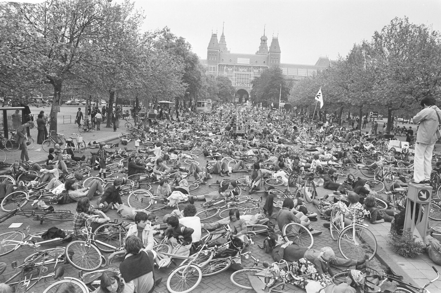 Eine schwarz-weiße Aufnahme einer Fahrraddemonstration mit zahlreichen Menschen, die mit ihren Rädern auf dem Boden liegen. Im Hintergrund stehen Bäume und ein historisches Gebäude mit Türmen.