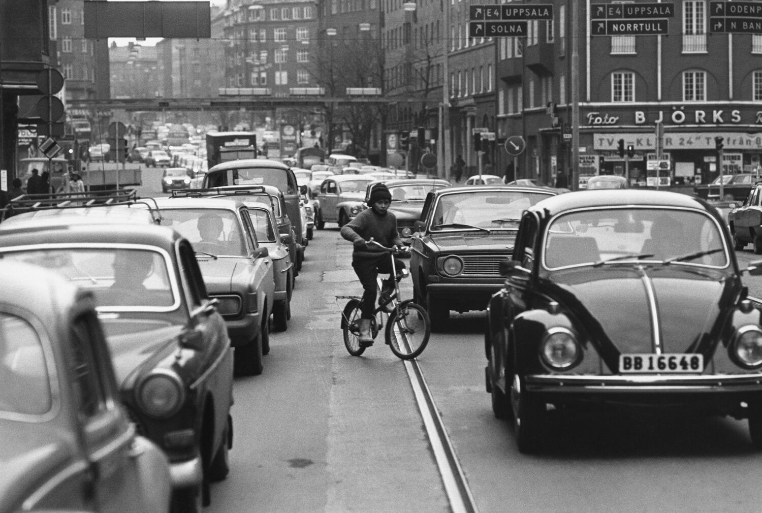 Ein Schwarz-Weiß-Foto einer belebten Stadtstraße mit dichter Autokolonne. Ein junger Mann fährt auf einem Fahrrad zwischen den Autos, umgeben von hohen Gebäuden und Straßenschildern.