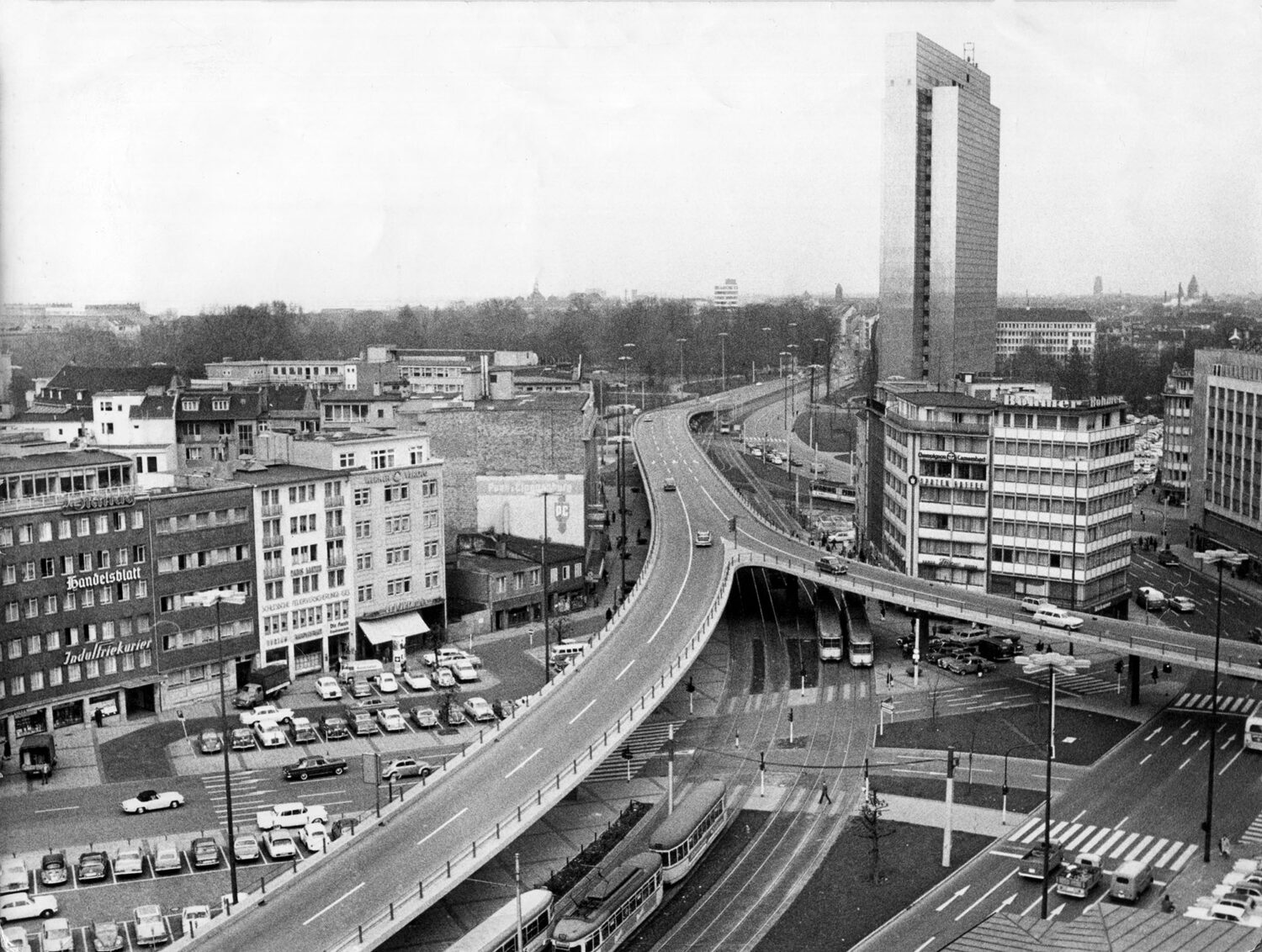 Stadtansicht in Schwarz-Weiß mit Hochhäusern, einer erhöhten Straßenbrücke, Straßenbahngleisen und geparkten Autos. Im Vordergrund mehrere Geschäftsgebäude mit sichtbarer Werbung.