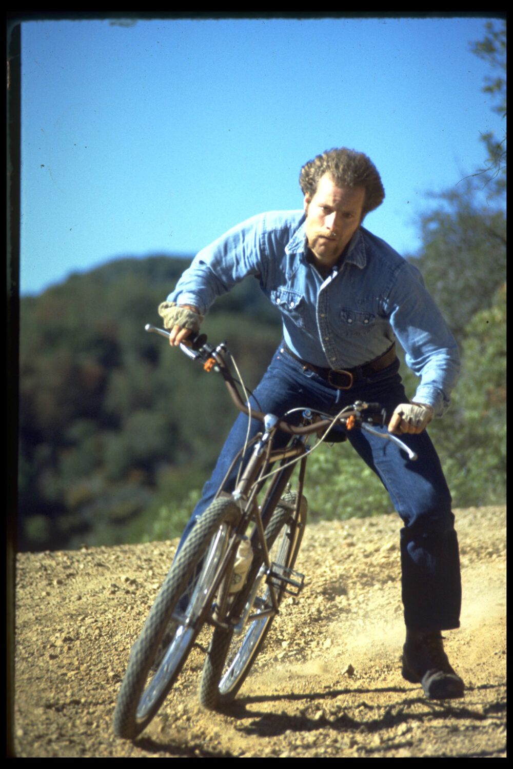 Ein Mann in Jeanshemd und Jeans fährt ein Mountainbike auf einem staubigen Weg durch eine hügelige Landschaft. Der Himmel ist klar und blau, und Bäume sind im Hintergrund sichtbar.