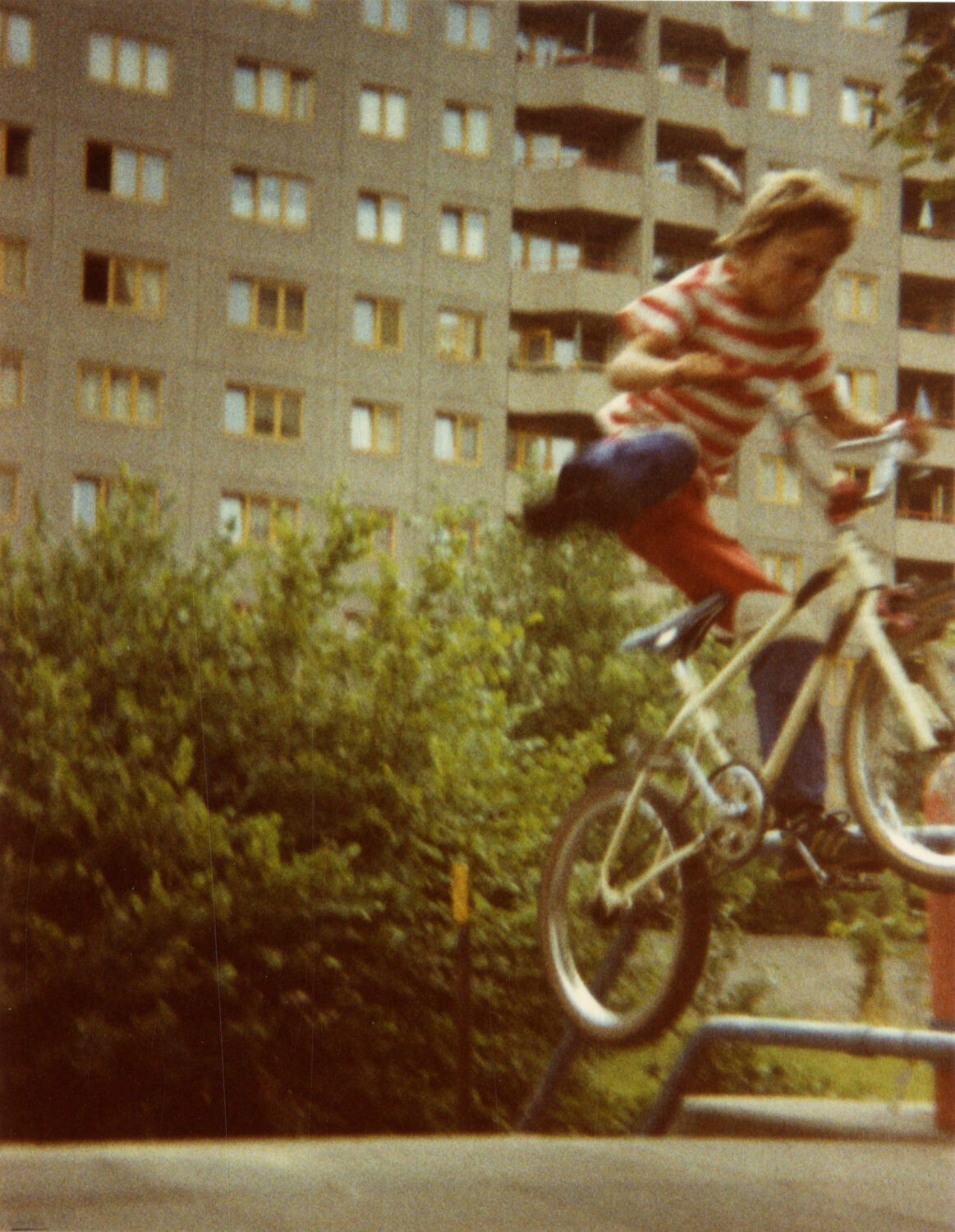 Ein Kind in rot-weiß gestreiftem T-Shirt und roter Hose springt mit einem weißen BMX-Rad über eine Stange, im Hintergrund sind Bäume und ein graues Wohngebäude mit vielen Fenstern zu sehen.