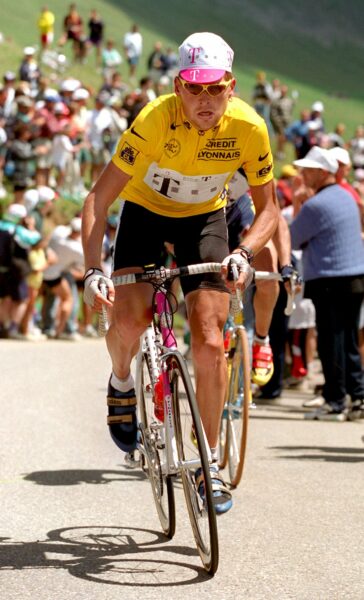 Ein Radrennfahrer in einem gelben Trikot fährt eine Steigung hinauf, umgeben von jubelnden Zuschauern. Der Sportler trägt eine weiße Mütze mit pinken Akzenten und gelbe Sonnenbrillen.