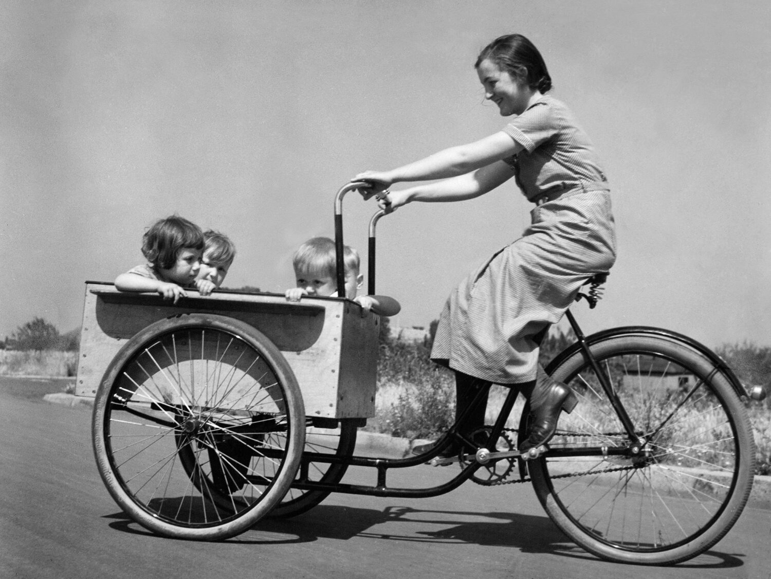 Eine Frau fährt ein Lastenfahrrad mit drei kleinen Kindern, die in der vorderen Holzkiste sitzen. Das schwarz-weiße Bild zeigt eine fröhliche Szene in einer ländlichen Umgebung.