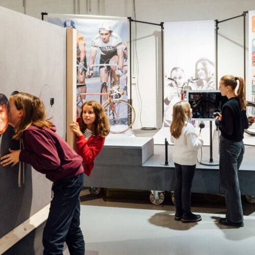 Drei Kinder und eine junge Frau erleben interaktive Stationen in einer Fahrrad-Ausstellung. Zwei Kinder untersuchen eine Wand mit Bildern, während die anderen beiden Personen an einem Bildschirm mit Mikrofonen interagieren. Im Hintergrund sind Fahrräder und große Poster zu sehen.