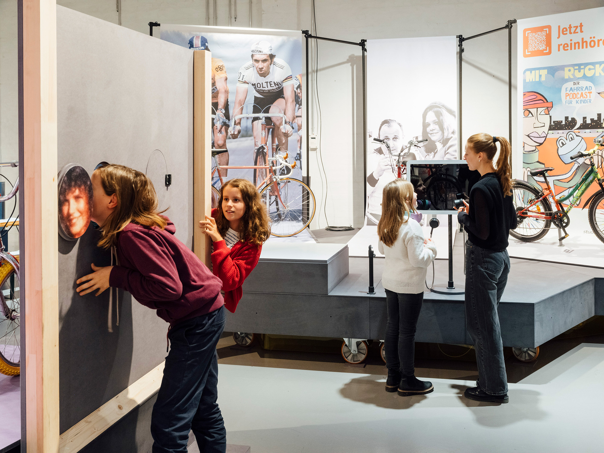 Drei Kinder und eine junge Frau erleben interaktive Stationen in einer Fahrrad-Ausstellung. Zwei Kinder untersuchen eine Wand mit Bildern, während die anderen beiden Personen an einem Bildschirm mit Mikrofonen interagieren. Im Hintergrund sind Fahrräder und große Poster zu sehen.