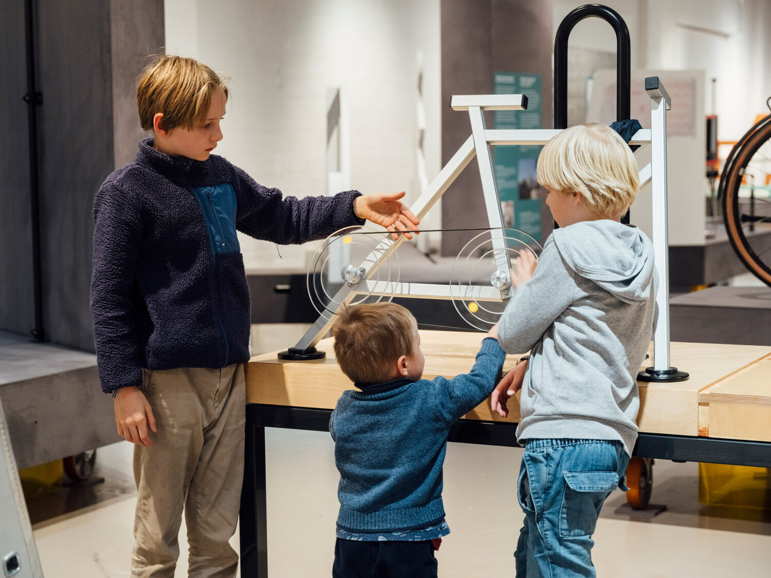 Drei Kinder interagieren mit einem weißen Modell eines Fahrrads in einer Ausstellung. Ein Junge zeigt auf die Pedale, während die anderen beiden neugierig zuschauen und die Details des Modells erkunden.