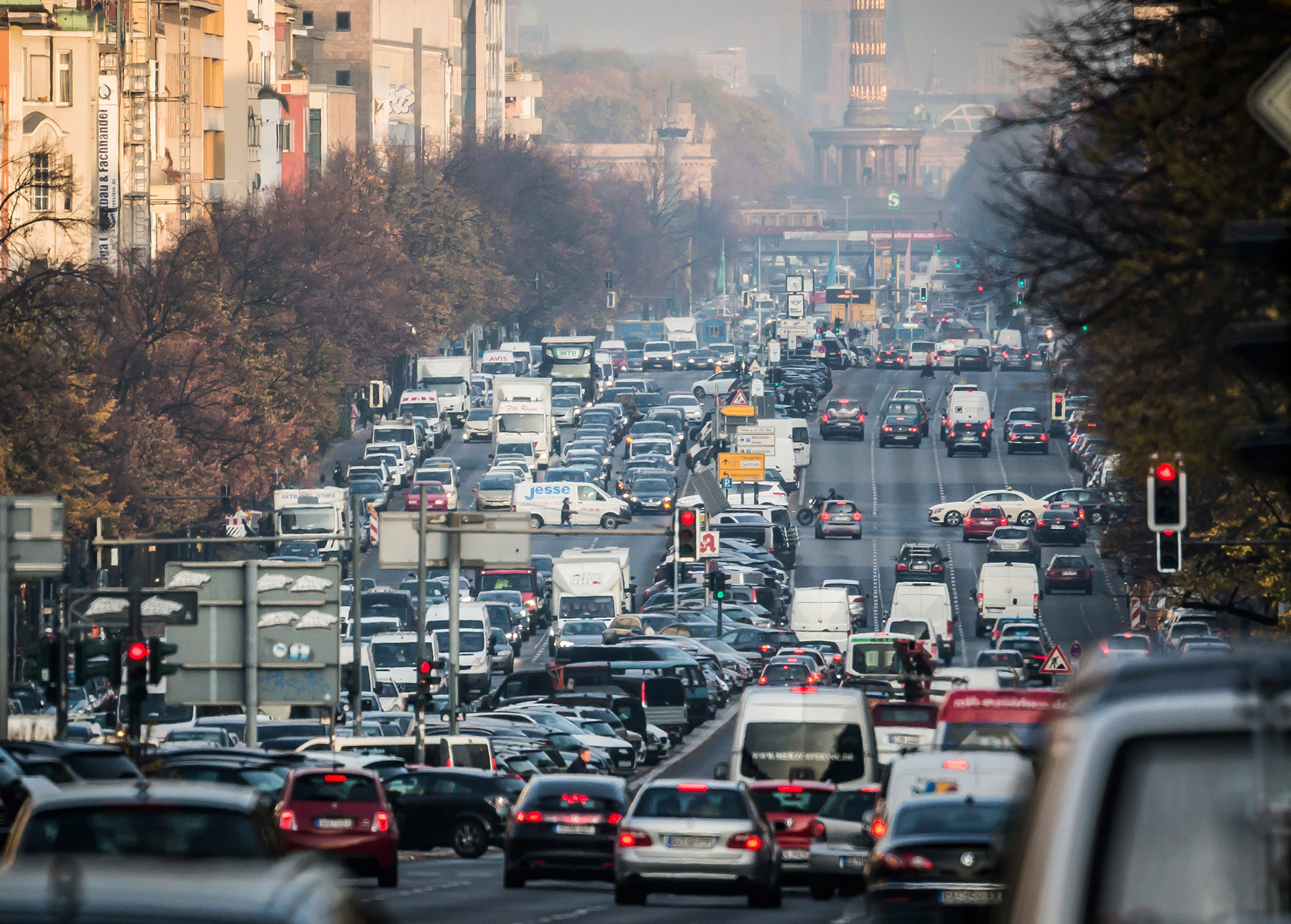 Dichter Autoverkehr auf einer breiten Straße in einer Großstadt. Zahlreiche Fahrzeuge stehen im Stau, im Hintergrund sind Bäume und eine Säule sichtbar, die eine städtische Landmarke darstellen könnte.