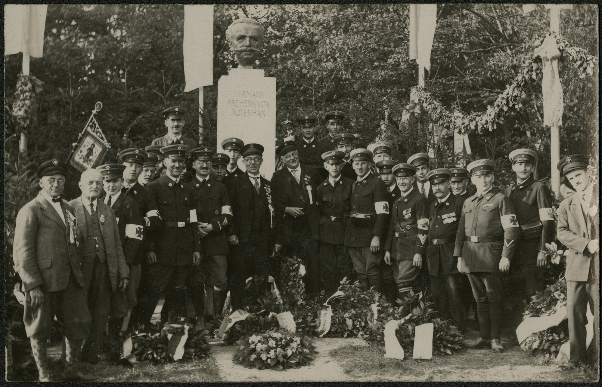 Schwarzweiß-Gruppenfoto vor einer Büste mit der Aufschrift „Hermann Freiherr von Rotenhan“: Mehrere Männer in Uniformen tragen Armbinden mit Kreuzsymbol, links steht eine Fahne. Im Vordergrund liegen Kränze.