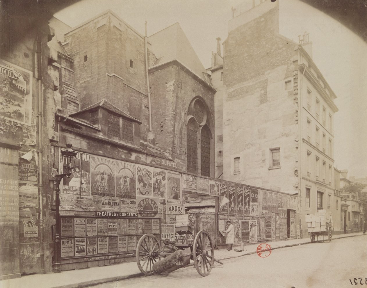 Historische Straßenansicht in Paris: An einer Mauer neben einer Kirche sind dichte Werbeplakate zu sehen. Ein Holzhandkarren steht davor, umliegende Gebäude ragen in den Hintergrund.