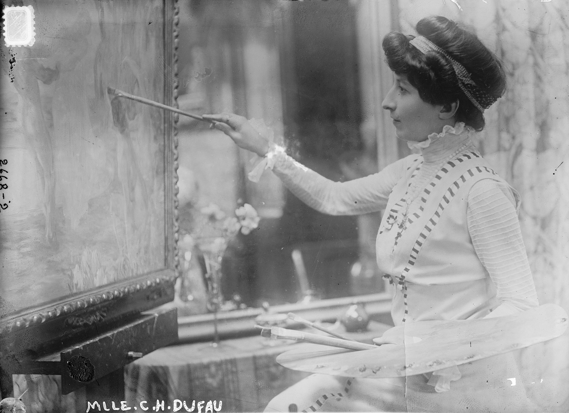 Schwarzweißfoto einer Frau mit hochgestecktem Haar, die an einer Staffelei malt und eine Palette hält. Am unteren Bildrand ist der Schriftzug „Mlle. C.H. Dufau“ zu sehen.