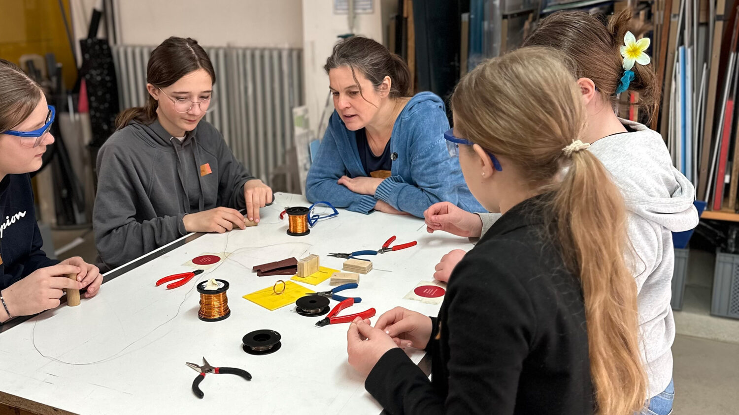 Fünf Personen arbeiten gemeinsam an einem Tisch in einer Werkstatt. Diverse Werkzeuge und Materialien wie Zangen und Drähte liegen auf dem Tisch.