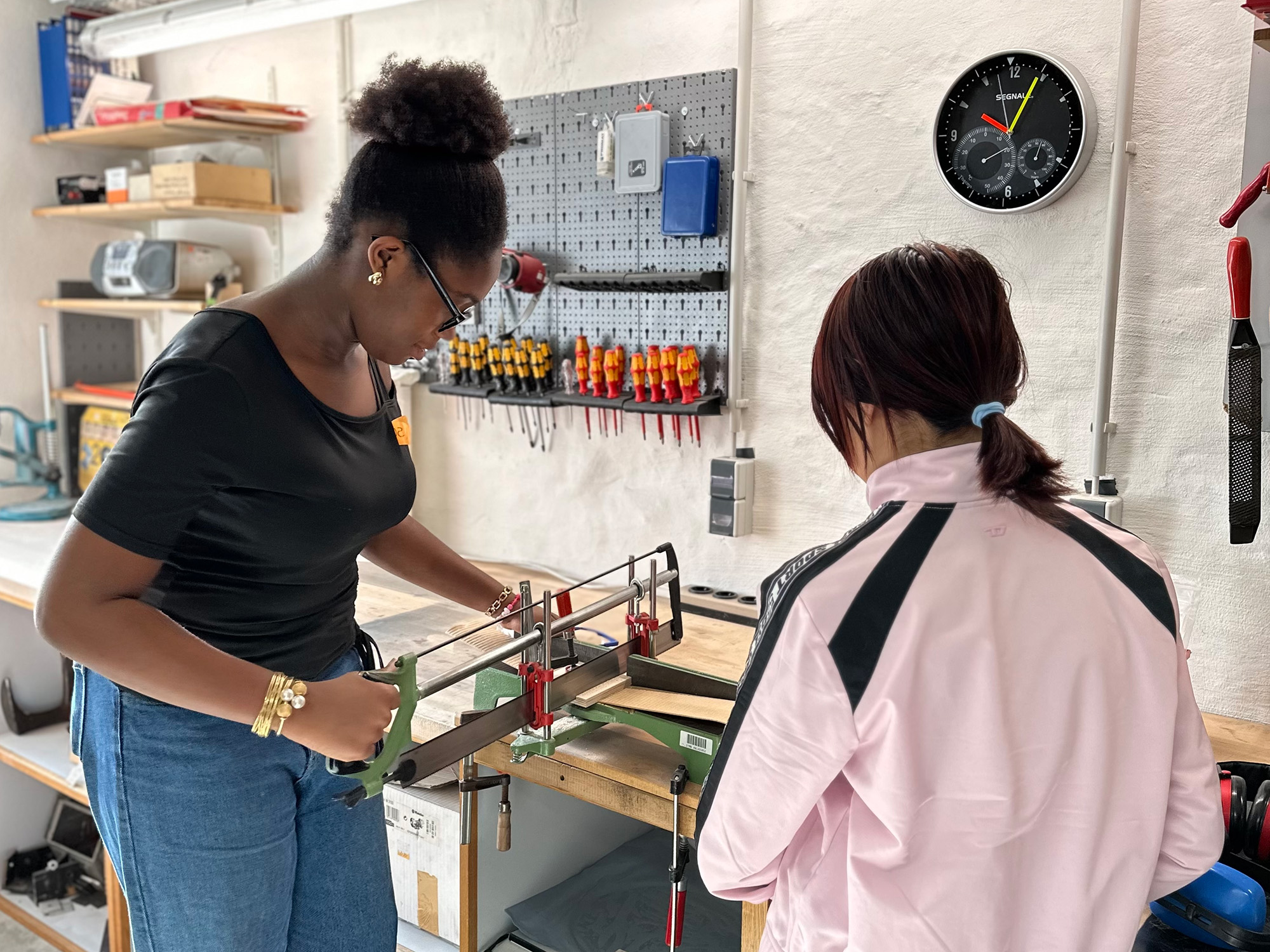 Zwei junge Frauen arbeiten in einer Werkstatt an einer Werkbank mit Werkzeugen und einer Bügelsäge. Eine Wanduhr und Regale mit Werkzeugen im Hintergrund.