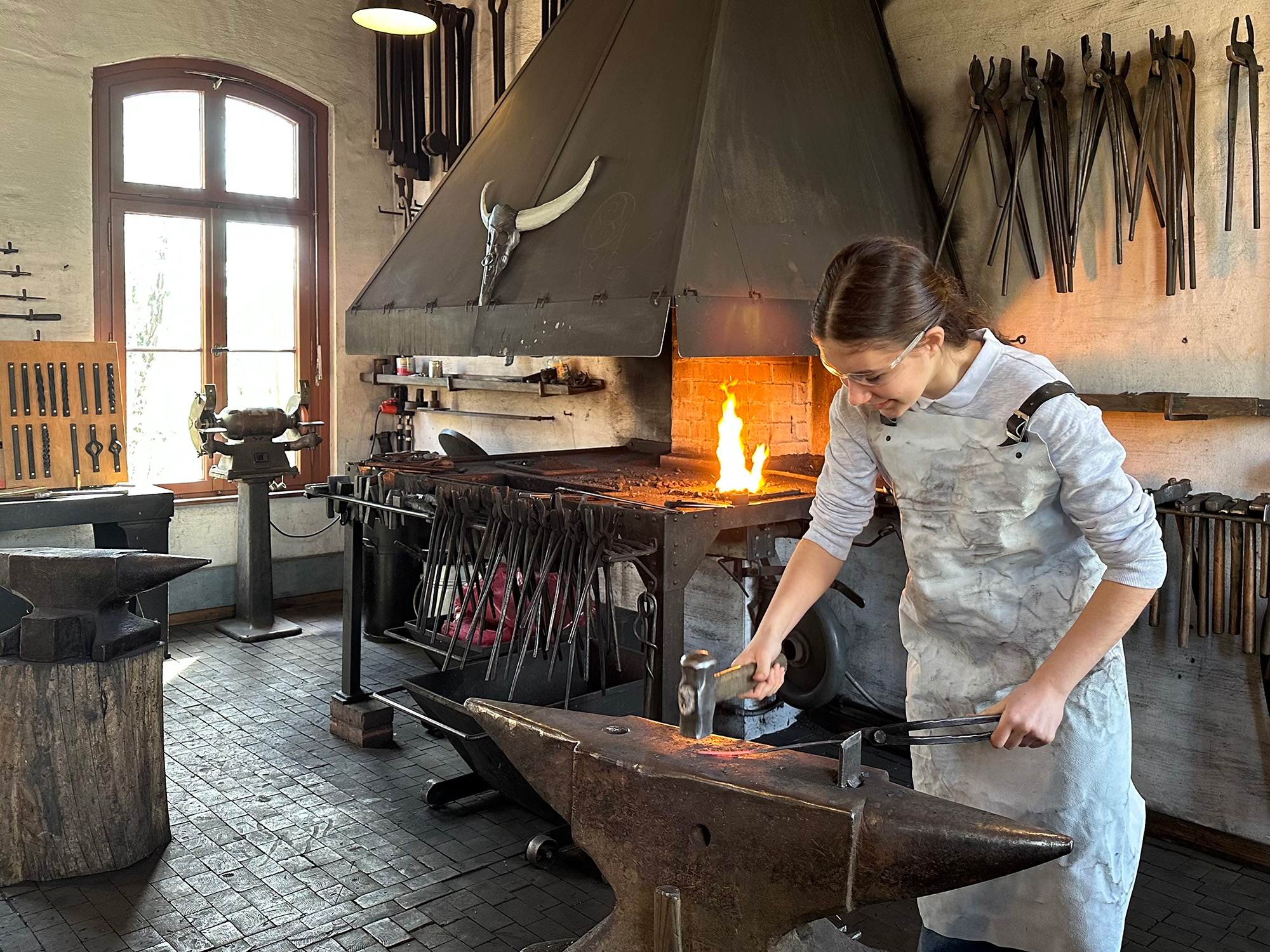 Eine junge Frau in Schutzkleidung bearbeitet Metall an einem Amboss in einer Schmiede mit offenem Feuer und Werkzeugen im Hintergrund.
