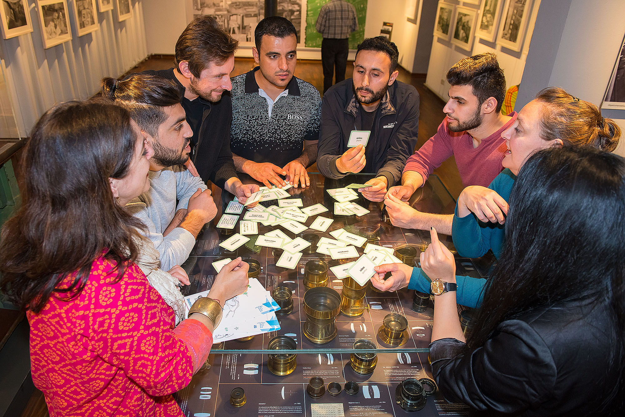 Eine Gruppe von Personen diskutiert an einer Vitrine mit historischen Objektiven in einem Museum. Auf der Vitrine liegen Kärtchen.