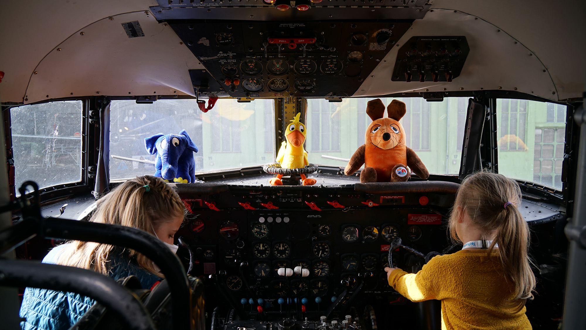 Zwei Kinder sitzen im Cockpit eines Flugzeugs mit Stofftieren auf dem Armaturenbrett.