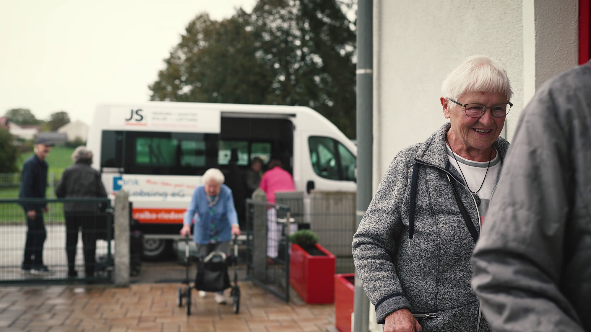 Eine ältere Frau mit Brille und grauer Jacke steht lächelnd im Vordergrund, während andere Personen aus einem Bus aussteigen.