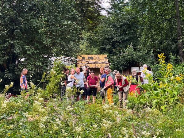 Eine Gruppe von Personen steht in einem üppigen Garten und betrachtet Pflanzen. Im Hintergrund befindet sich eine kleine Holzhütte.