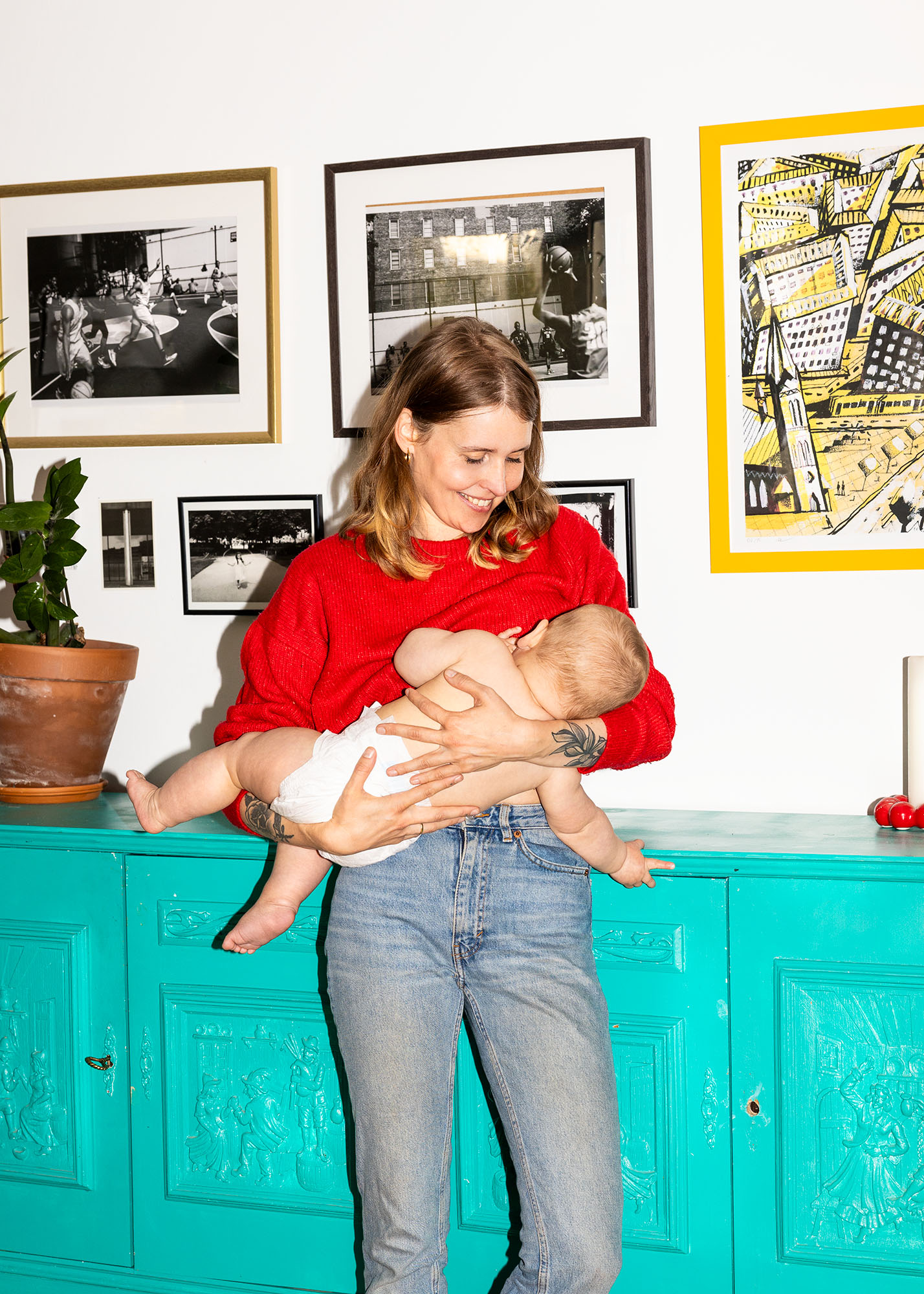 Eine Frau in einem roten Pullover stillt ein Baby und steht vor einem türkisfarbenen Schrank mit Bildern an der Wand.