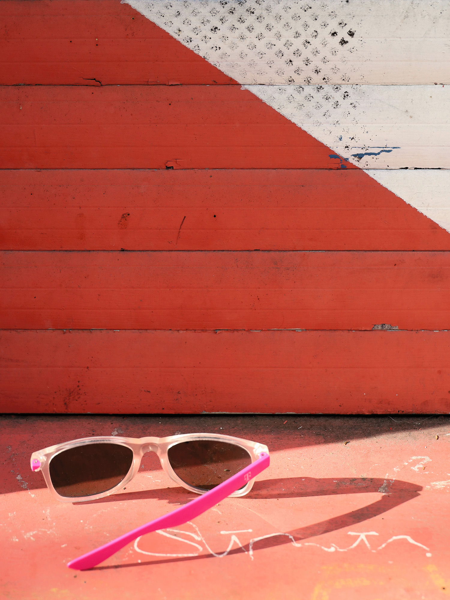 Eine kaputte Sonnenbrille mit pinkfarbenen Bügeln liegt auf dem Boden vor einer roten und weißen Wand.