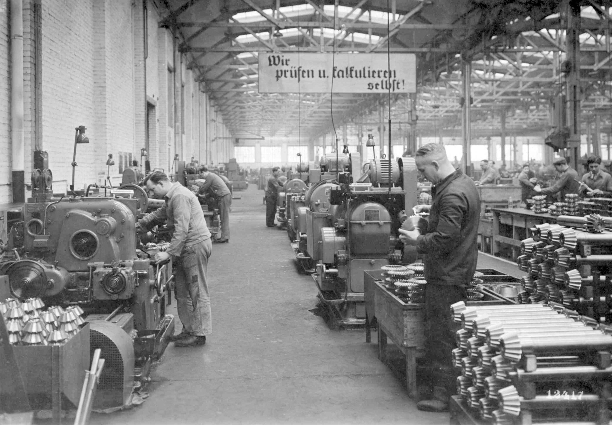 Arbeiter in einer historischen Fabrikhalle mit Maschinen und einem Banner, das "Wir prüfen und kalkulieren selbst!" zeigt.