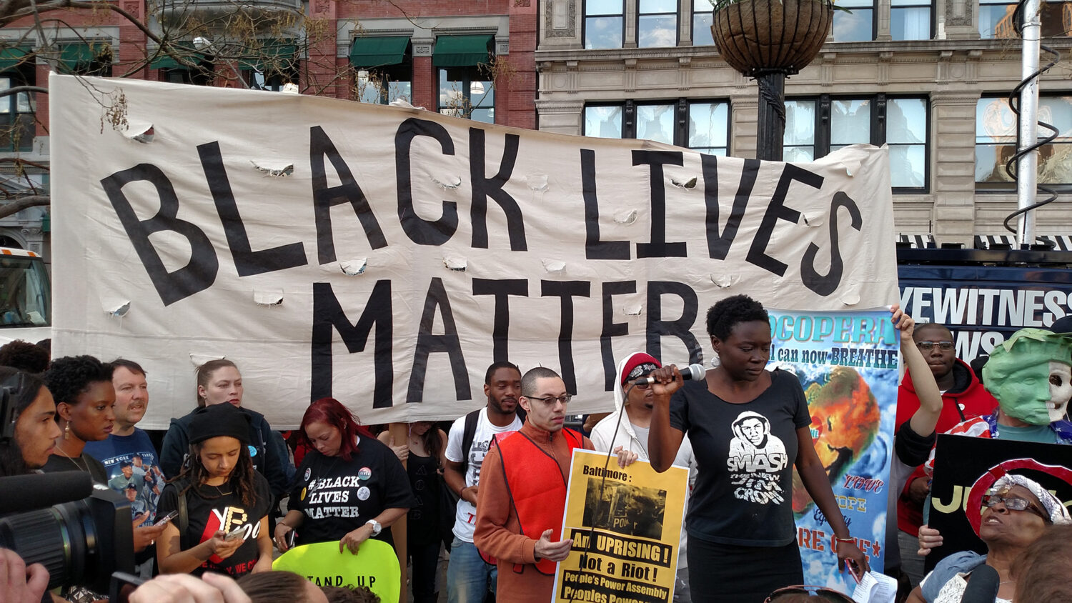 Eine Gruppe von Demonstranten hält ein großes Banner mit der Aufschrift "Black Lives Matter" und weitere Plakate in einer Stadt.