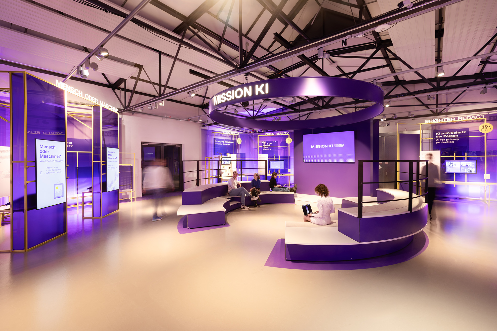 Eine moderne Ausstellung mit dem Thema "Mission KI" zeigt verschiedene interaktive Stationen und Sitzbereiche in einem lila beleuchteten Raum.