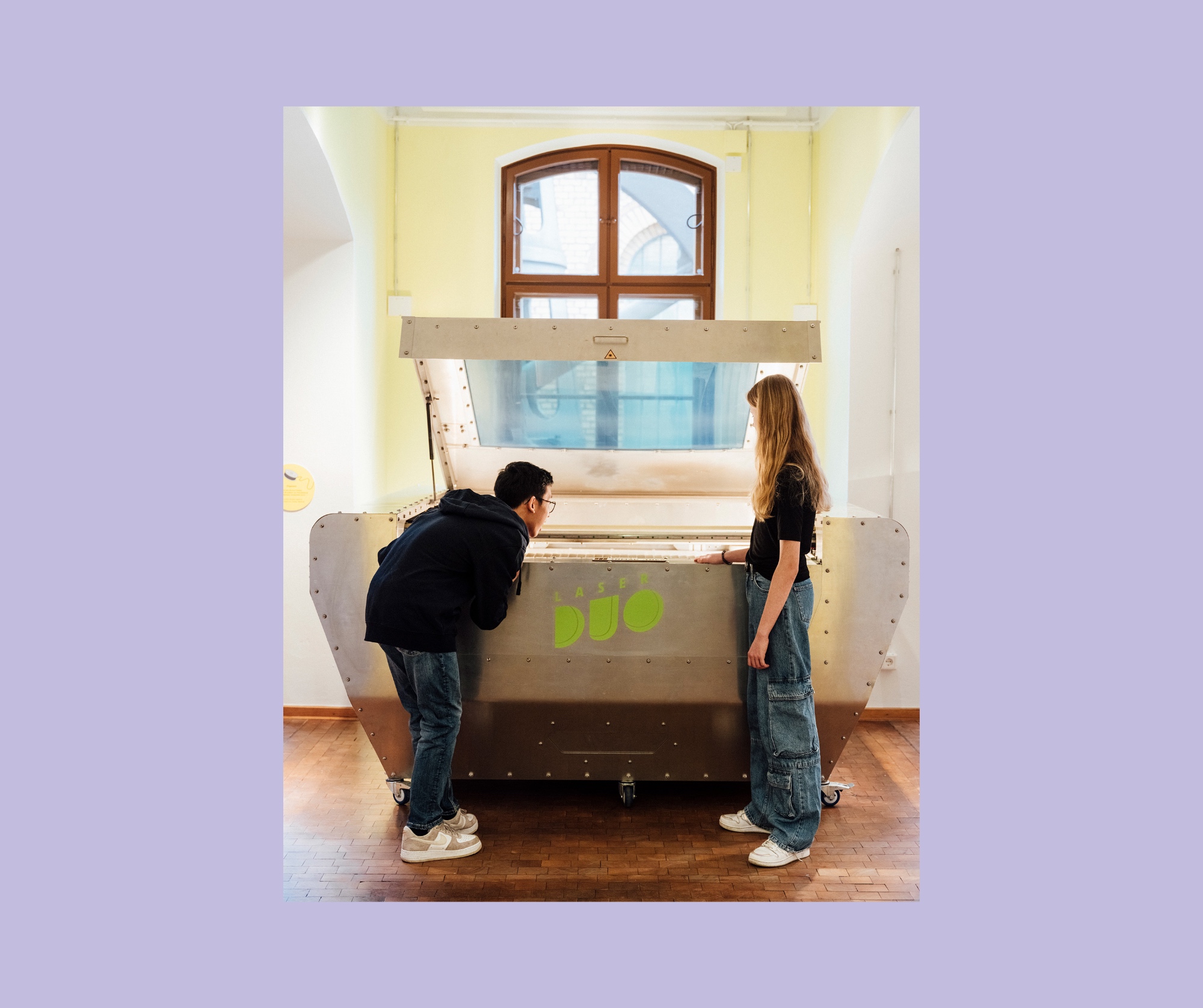 Zwei Personen betrachten eine große, metallische Maschine mit der Aufschrift "Laser Duo" in einem Raum mit großen Fenstern.