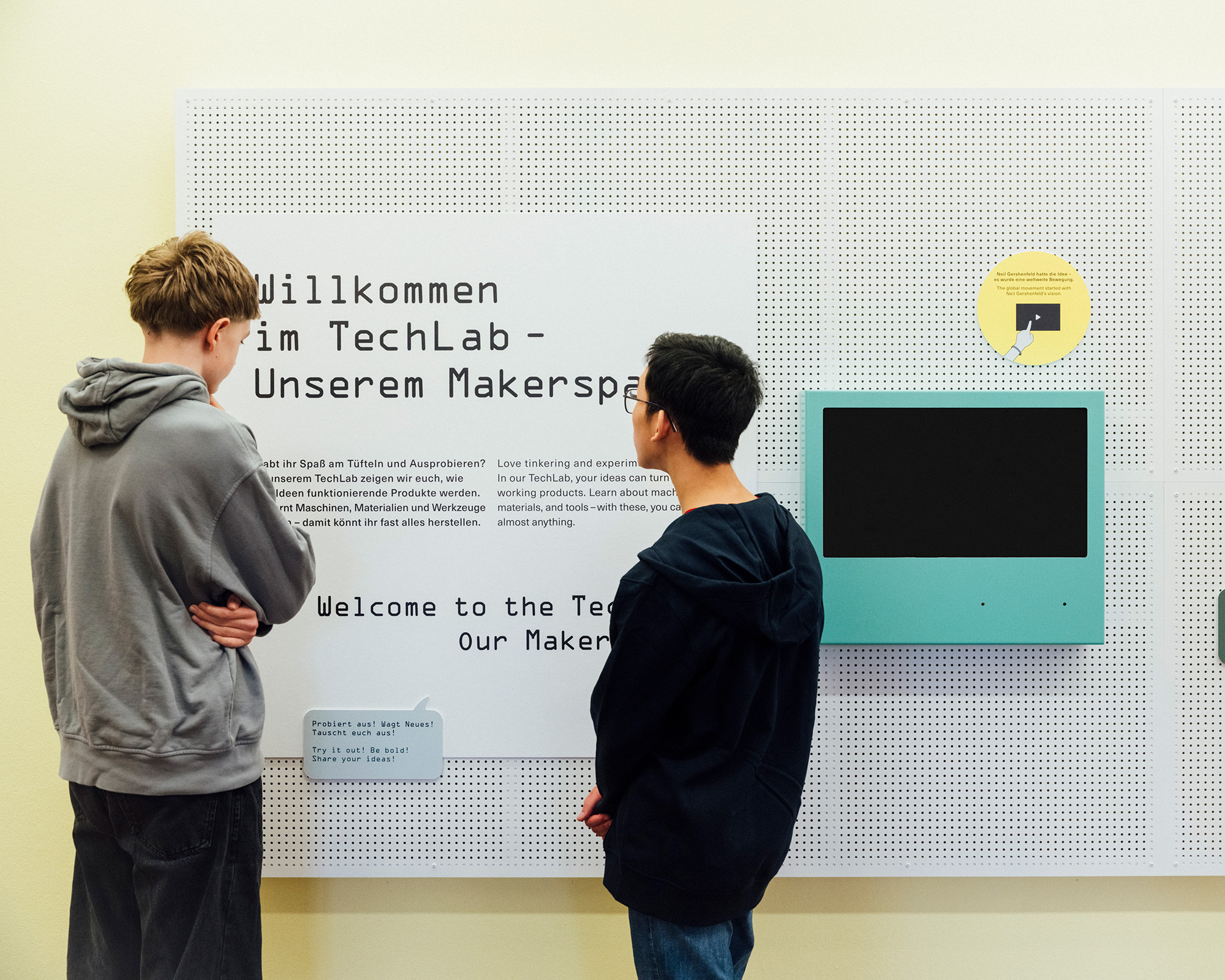 Zwei Personen stehen vor einer Tafel mit der Aufschrift "Willkommen im TechLab - Unserem Makerspace" und einem Bildschirm.