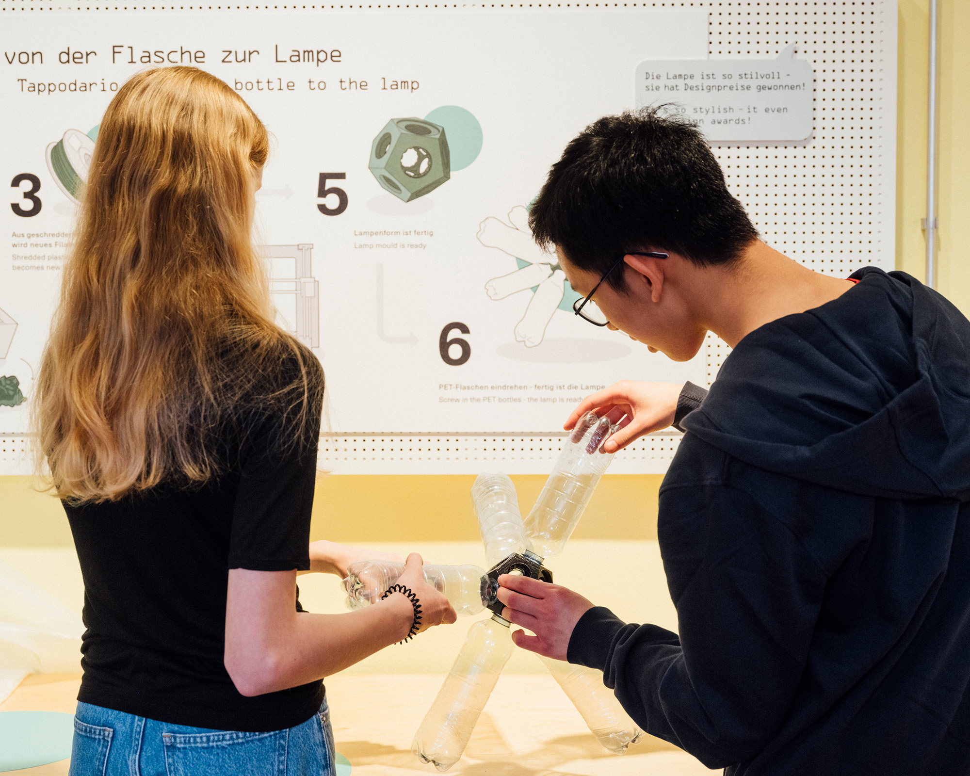 Zwei Personen arbeiten an einer Konstruktion aus Plastikflaschen und betrachten eine Anleitung an der Wand.