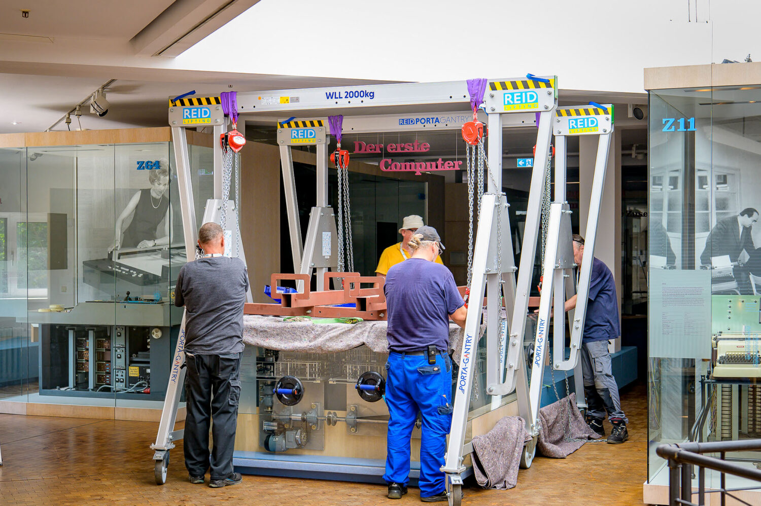 Vier Personen heben mit einem Kran eine große Glasvitrine vom Nachbau des historischen Computers Z1 in einer Ausstellung.