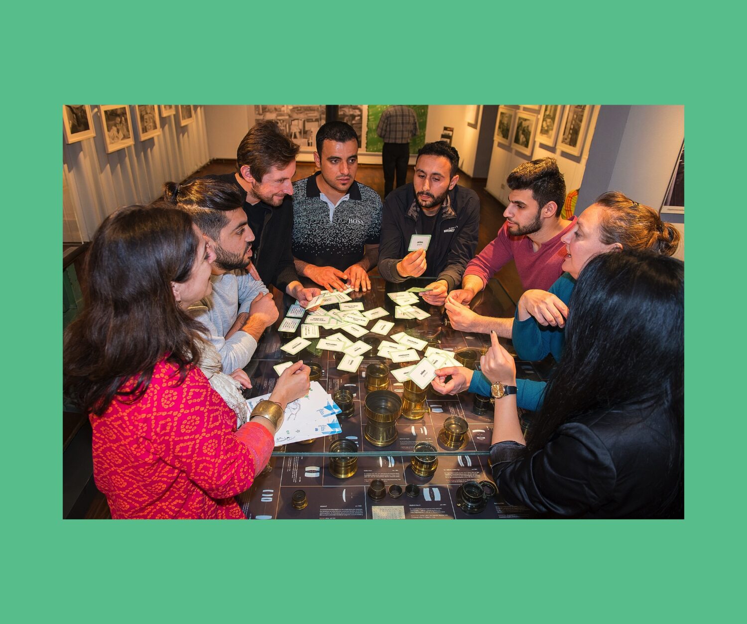 Eine Gruppe von acht Personen lernt mithilfe von Lernkarten Deutsch, an einer Vitrine mit historischen Fotoobjektiven in einem Museum.