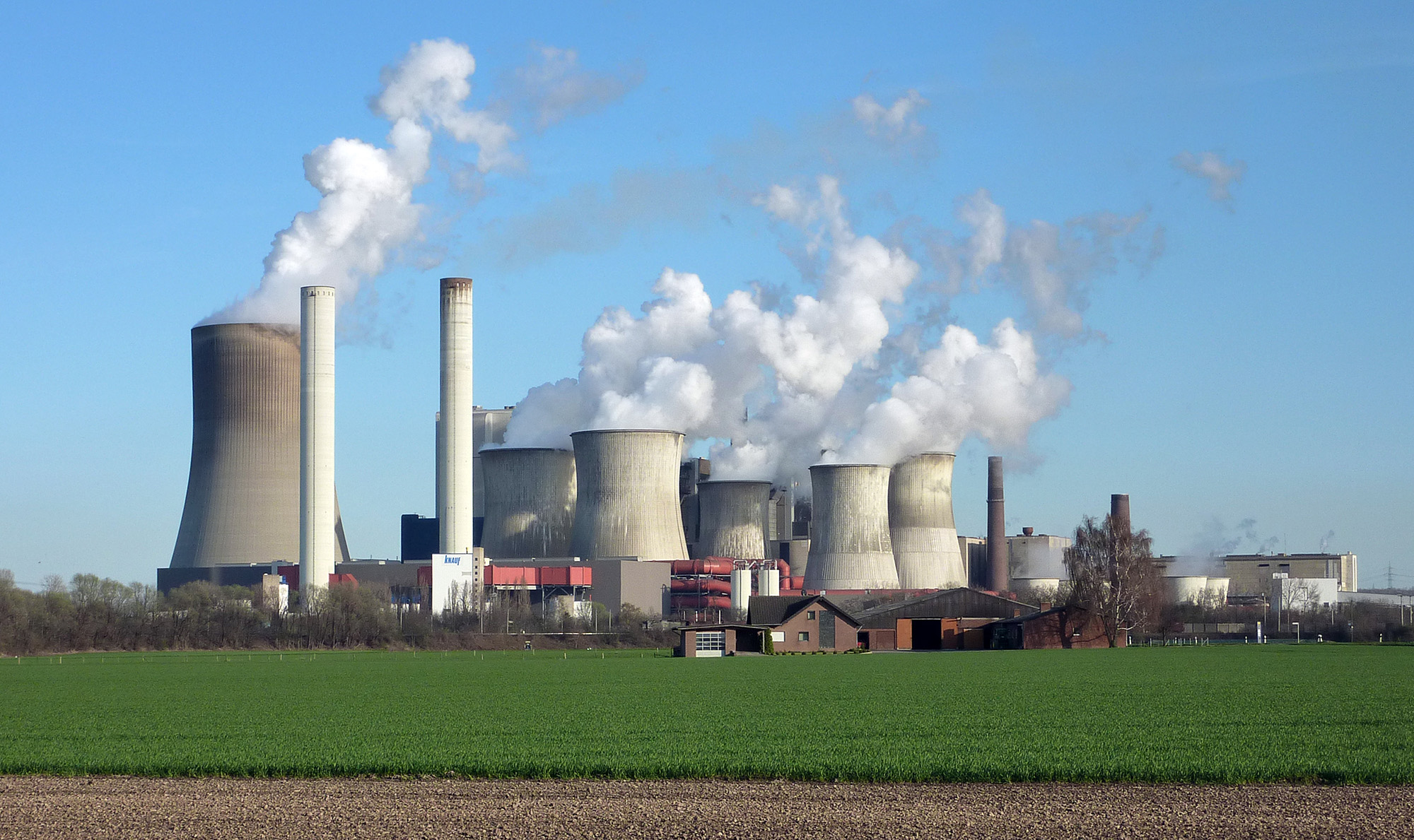 Großes Kohlekraftwerk mit mehreren rauchenden Kühltürmen und Industrieanlagen vor grünen Feldern und blauem Himmel.