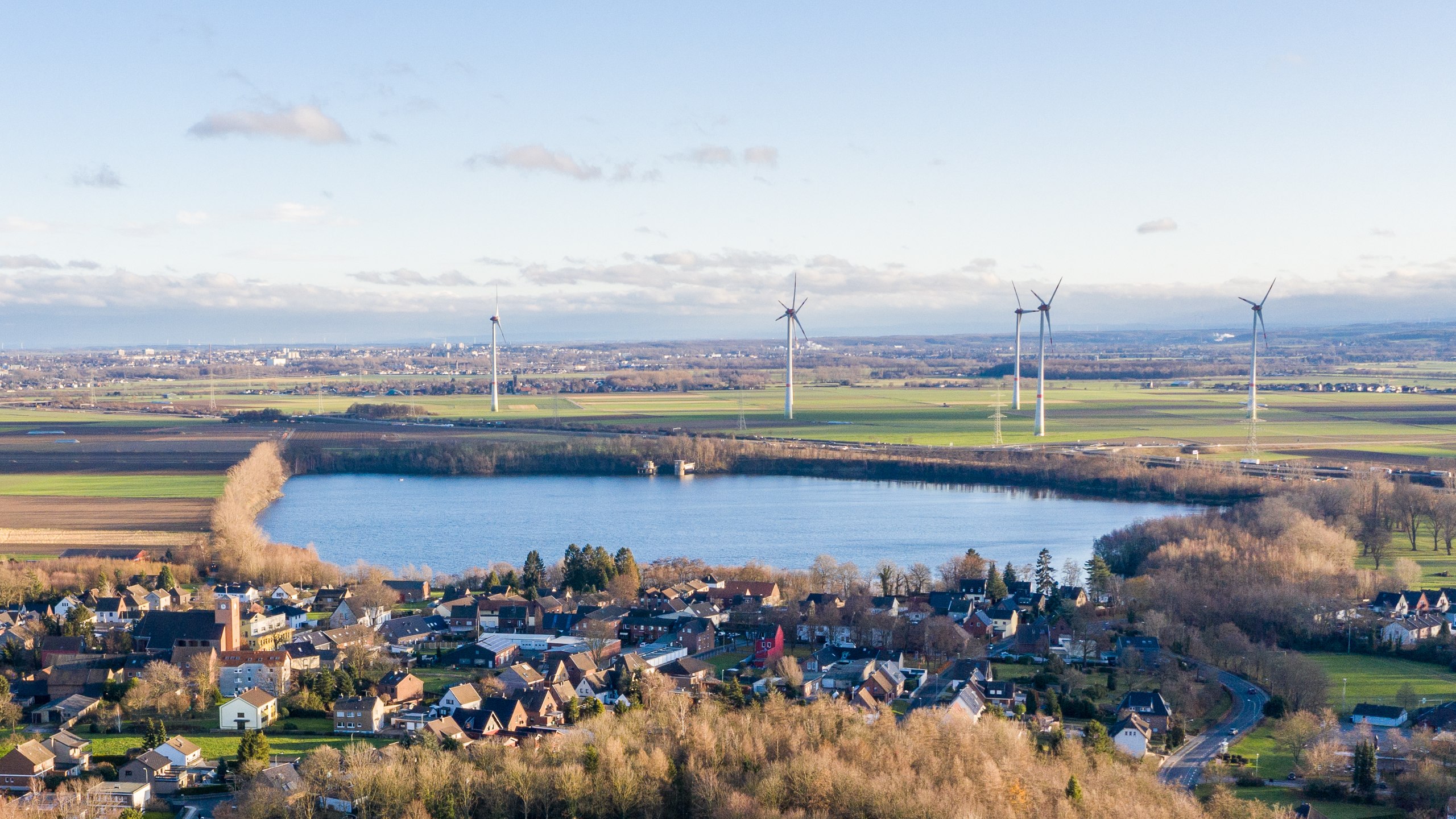 Dorf am Seeufer mit Windkraftanlagen im Hintergrund, umgeben von Feldern, Wäldern und sanftem Hügelland unter leicht bewölktem Himmel.