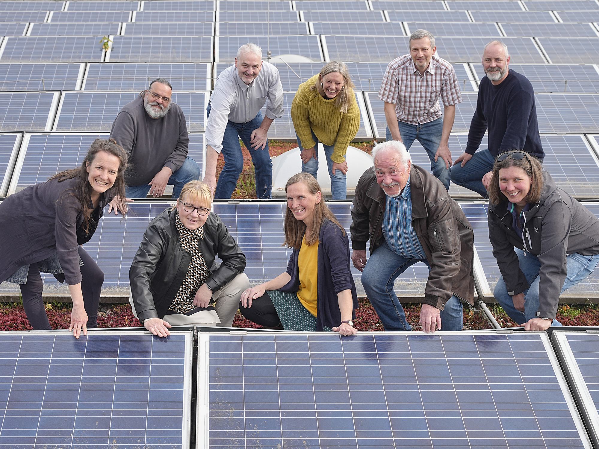 Gruppe von acht lächelnden Personen posiert auf einer Solaranlage, einige kniend und stehend, vor blauen Solarpanelen.