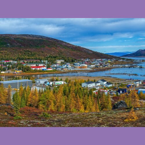 Kleinstadt am Meer mit bunten Häusern, Industriegebiet und Nadelwäldern, umgeben von Bergen und Küstenlandschaft unter bewölktem Himmel.