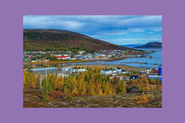 Kleinstadt am Meer mit bunten Häusern, Industriegebiet und Nadelwäldern, umgeben von Bergen und Küstenlandschaft unter bewölktem Himmel.