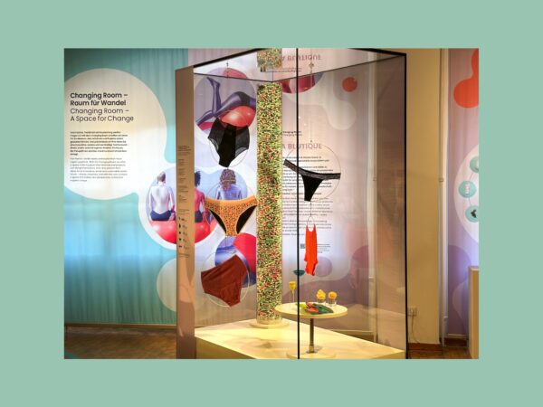 Ausstellung "Changing Room – Raum für Wandel": Vitrine mit hängenden Kleidungsstücken, Säule mit Tampons und Infotafeln zu nachhaltiger Mode.