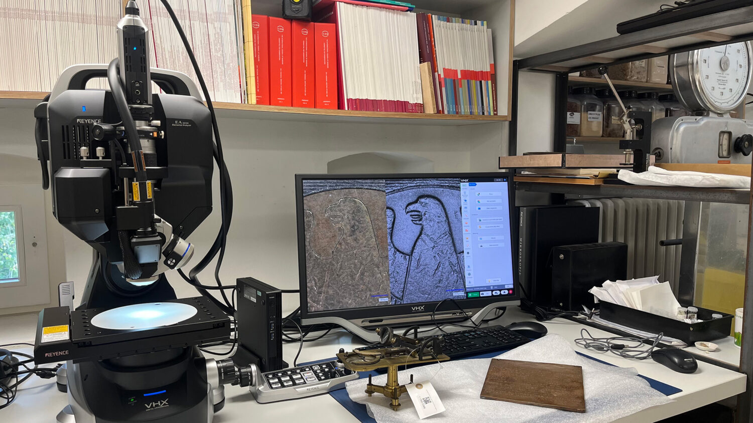 Digitales Mikroskop (Keyence VHX) in einem Labor mit Bildschirm, der eine detaillierte Aufnahme einer alten Gravur zeigt, umgeben von Büchern und Messgeräten.