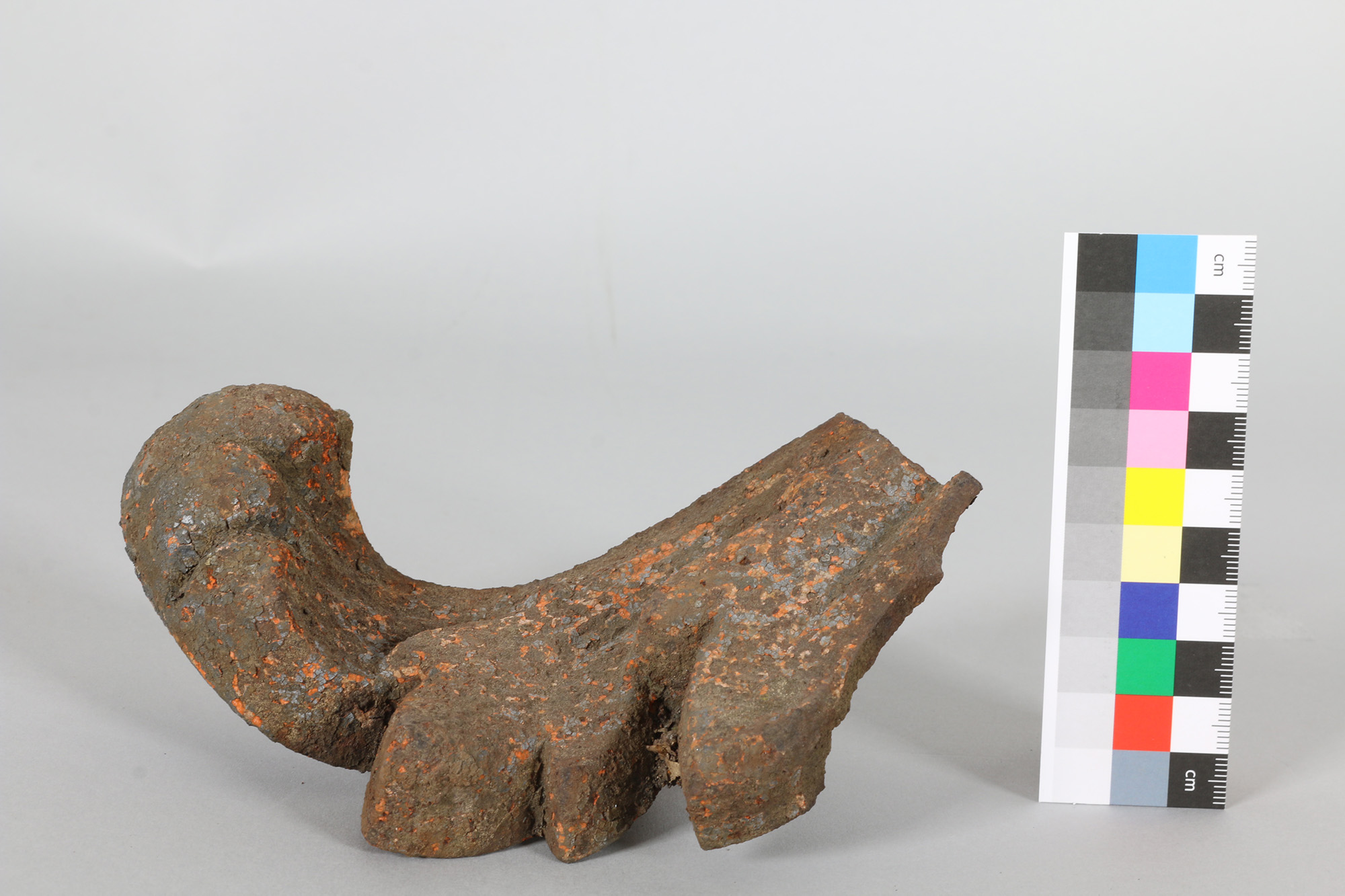 Stark korrodiertes, gebogenes Metallrohrstück mit Rostspuren, daneben ein Maßstab in Zentimetern und Farbskala zur Größenreferenz.