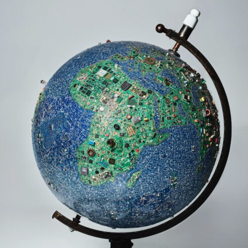 Globus aus unzähligen elektronischen Bauteilen und Platinen, die die Kontinente bilden, auf schwarzem Ständer vor hellem Hintergrund.