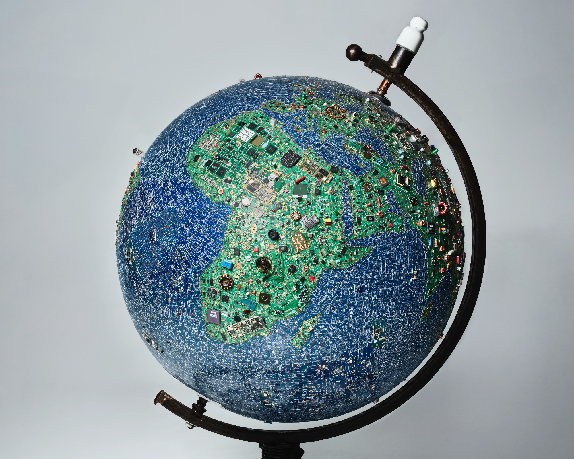 Globus aus unzähligen elektronischen Bauteilen und Platinen, die die Kontinente bilden, auf schwarzem Ständer vor hellem Hintergrund.