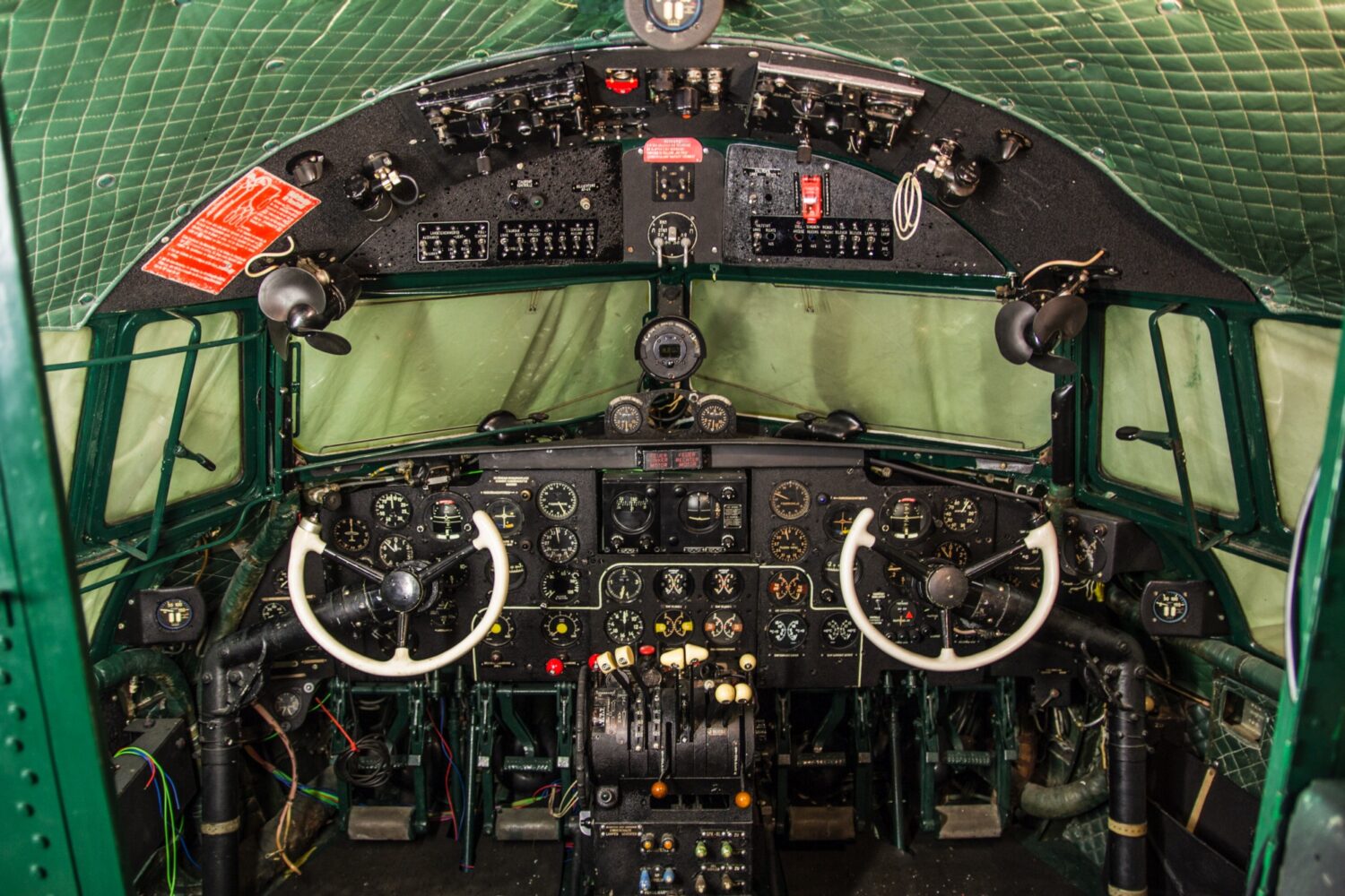 Restauriertes historisches Flugzeugcockpit mit doppelter Steuerung, vielen analogen Rundinstrumenten, Schaltern und weißen Steuerknüppeln.