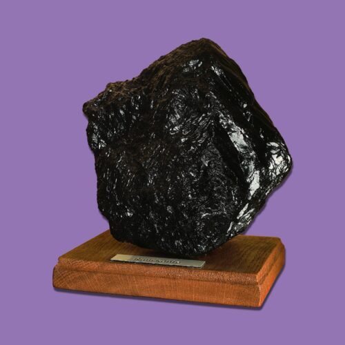 Großes Stück Anthrazitkohle auf einer Holzplinthe mit Beschriftung, vor violettem Hintergrund.
